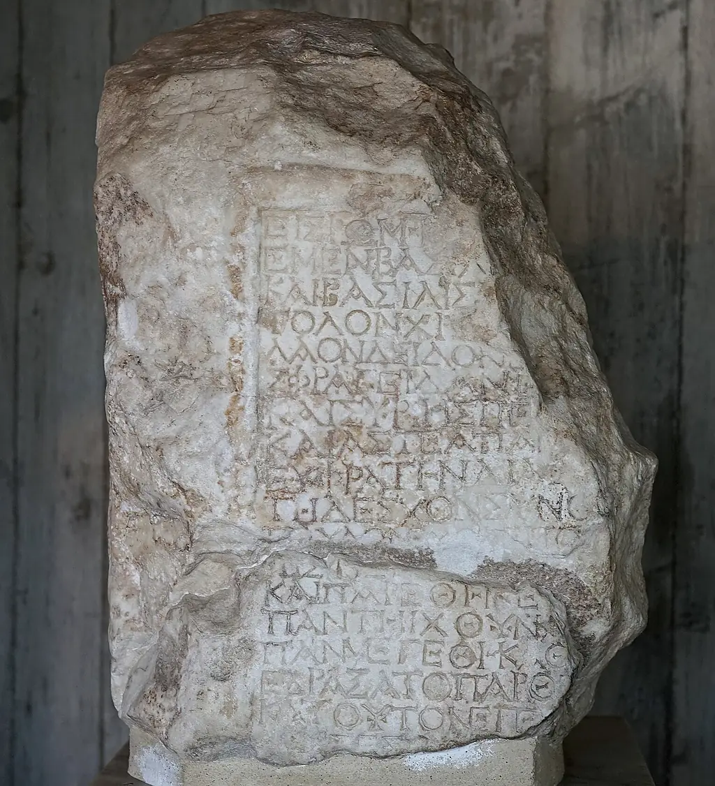 Das erhaltene Bruchstück der Grabinschrift: ein unregelmäßiger, heller Marmorblock mit eingemeißelten griechischen Großbuchstaben in zwei Feldern, auf einem Sockel vor einer Holzwand.
