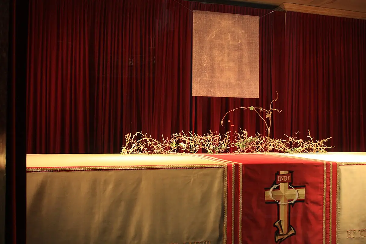 Vor einem roten Vorhang steht ein langer, verhüllter Schrein, belegt mit einem rot-weißen Tuch mit Kreuz und der Inschrift INRI; darauf liegen dornige Zweige, darüber hängt ein Bild des Gesichts vom Grabtuch.