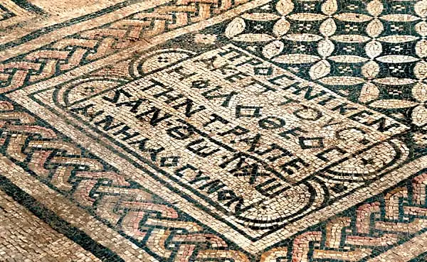 Mosaikboden mit geflochtenen Randbändern; in einem gerahmten Feld eine griechische Inschrift aus dunklen Steinchen auf hellem Grund, daneben ein Blattmuster.