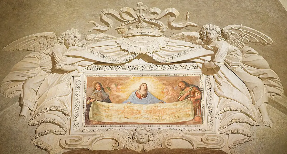 Ein Stuckrahmen mit zwei großen Engeln, darüber eine Krone; im gemalten Bild darin halten zwei Heilige das lange Grabtuch mit dem doppelten Bild des Mannes, hinter ihnen Maria im blauen Mantel und kleine Engelsköpfe.